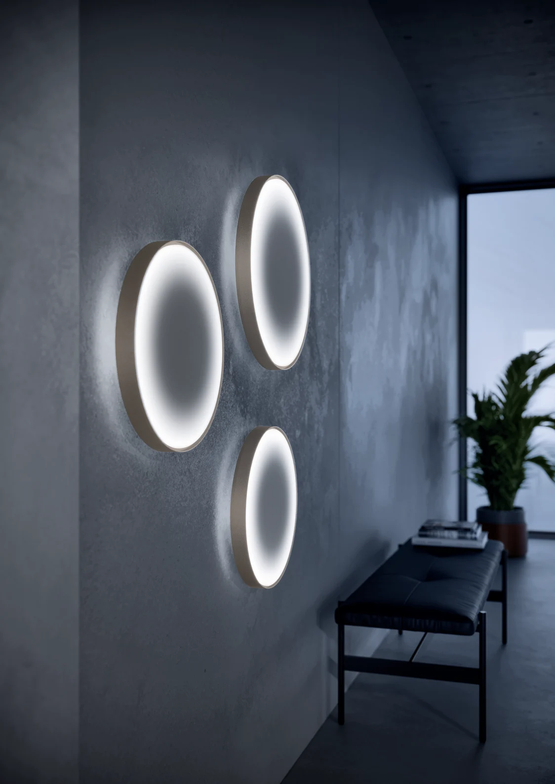 Trois appliques LED LANOS diffusent une lumière douce et homogène dans un couloir contemporain. À l’arrière-plan, on aperçoit un banc et une plante.
