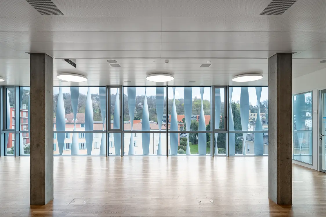 A light-filled, empty room with large window fronts and wooden flooring. Several concrete columns and round ceiling luminaires define the modern ambiance.