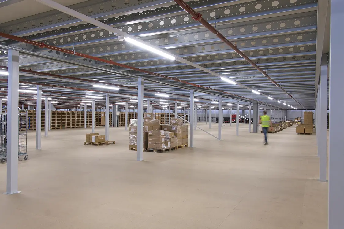 Grande capannone luminoso con pilastri e scaffali bianchi e con cartoni impilati su pallet. Un dipendente con giubbino ad alta visibilità attraversa l'ambiente.