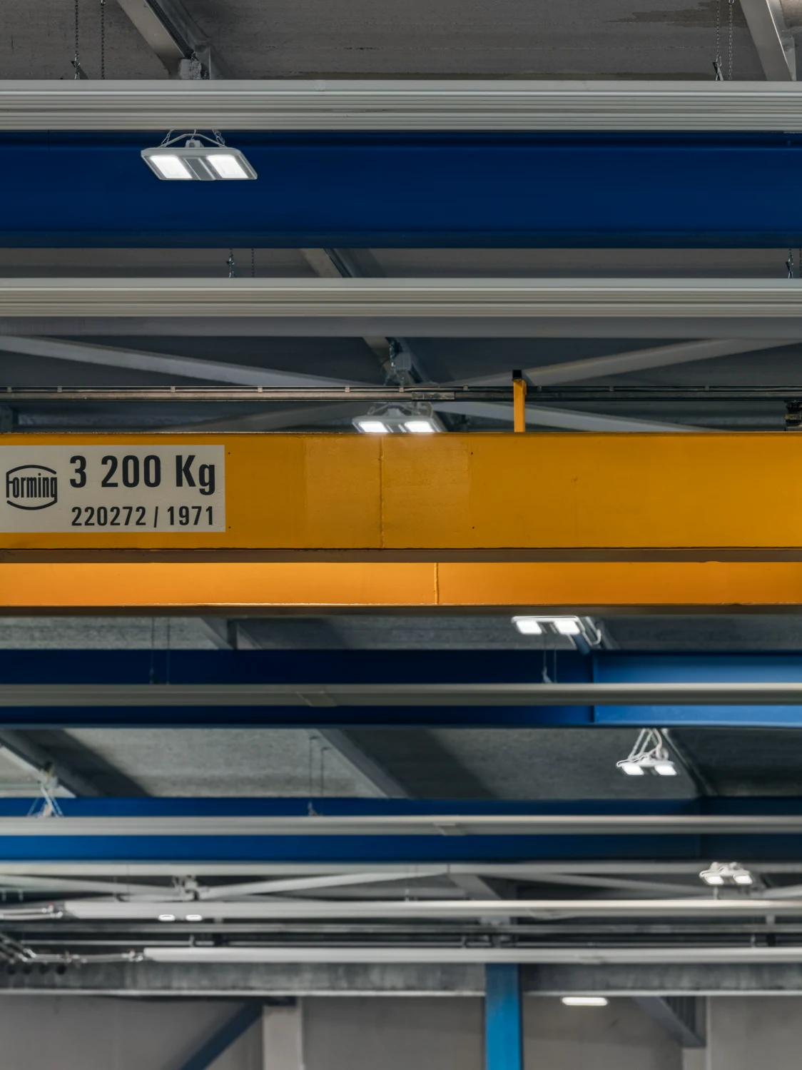 Industriehalle mit blauem Stahlträger und gelbem Brückenkran, ergänzt durch eine moderne, hängende LED-Leuchte. Die effiziente Beleuchtung sorgt für optimale Sichtverhältnisse in der Arbeitsumgebung.
