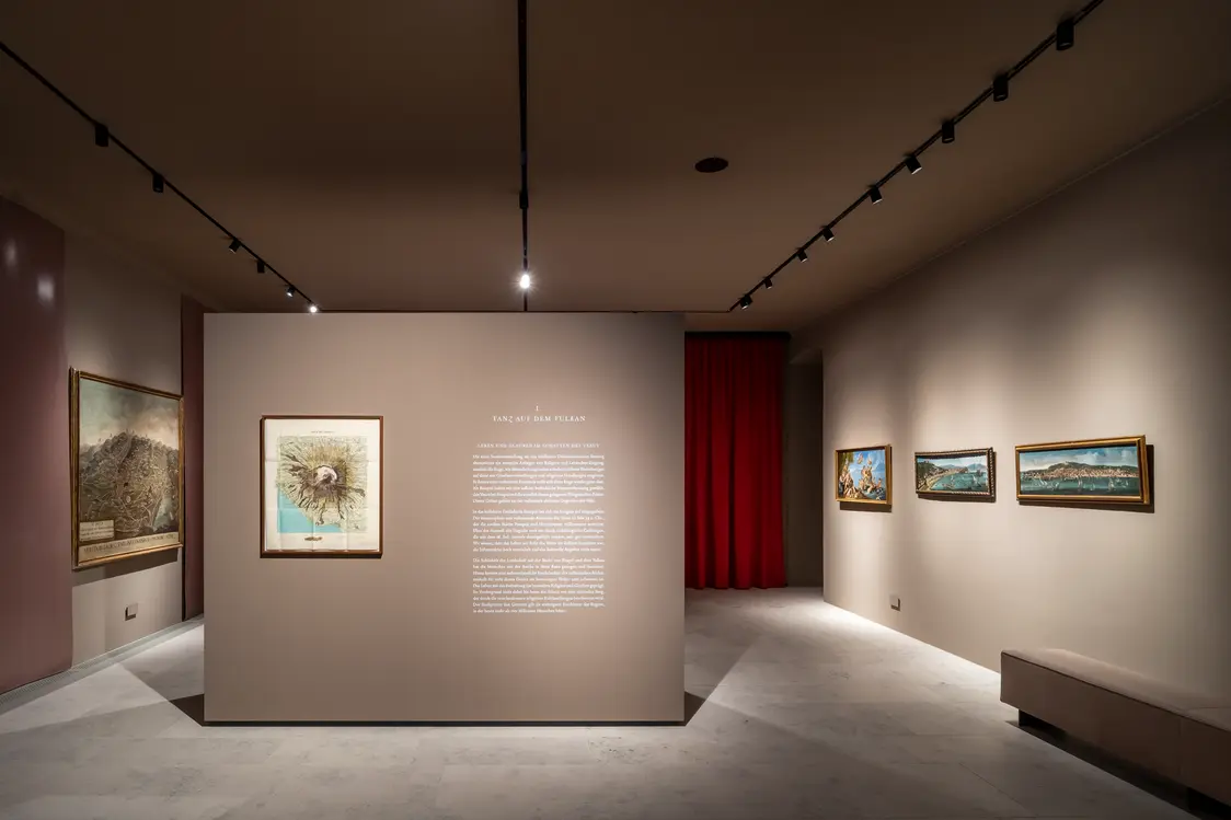 Ein Ausstellungsraum mit neutralen Wänden, in dem gezielte Strahler die Kunstwerke und die zentrale Infotafel hervorheben. Die gerichtete Beleuchtung schafft Fokus und hebt Details hervor.