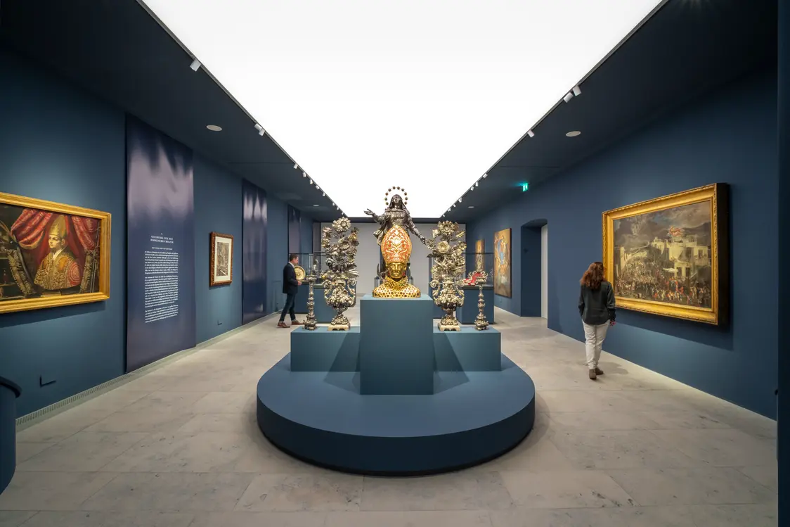 Eine Galerie mit blauen Wänden, in der eine Skulptur und Gemälde durch helles, diffuses Deckenlicht und zusätzliche Spotlights optimal inszeniert werden. Die Beleuchtung sorgt für eine klare, freundliche Wirkung und lenkt den Blick auf die Kunstwerke.