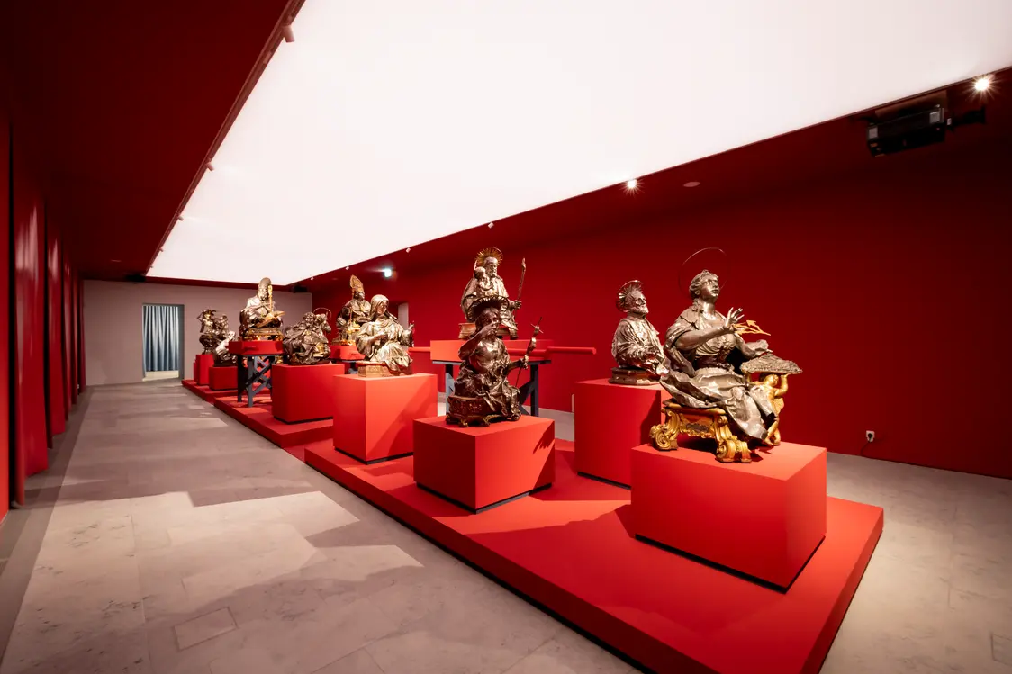 Ein Ausstellungsraum mit roten Wänden und Podesten, auf denen Skulpturen stehen, beleuchtet durch eine großflächige, helle Deckenbeleuchtung. Das gleichmäßige Licht hebt die Farben und Formen der Exponate besonders hervor.