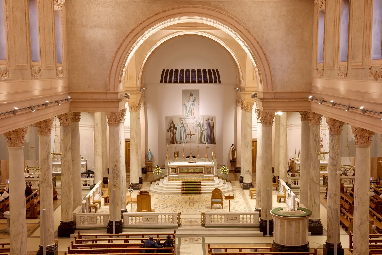 Catehdral of Saint Patrick and Saint Felim, Cavan - View of a cathedral sanctuary framed by tall marble columns and a large arched ceiling. A white marble altar stands at the center beneath religious artwork, with a baptismal font, wooden pews, and a few seated visitors visible in the foreground
