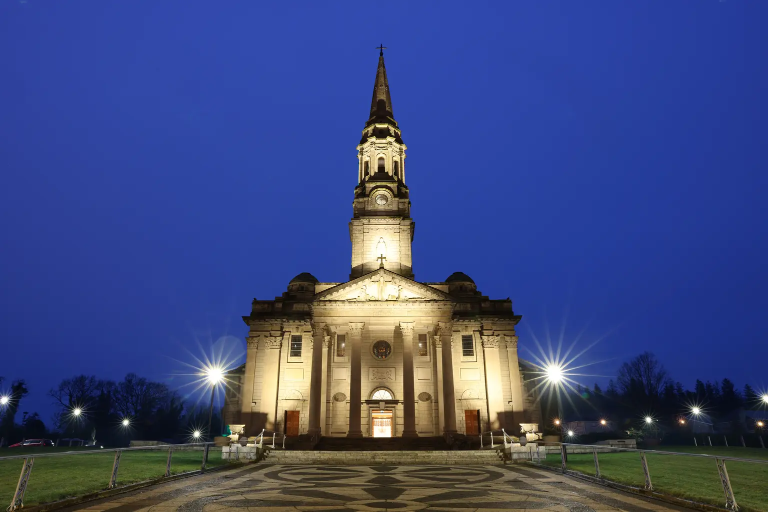 Cathedral of Saint Patrick and Saint Felim, Cavan - Illuminated cathedral with a tall central spire and grand columned entrance, photographed at dusk under a deep blue sky. A patterned stone walkway leads to the front steps, with glowing streetlights and grassy lawns framing the symmetrical scene.