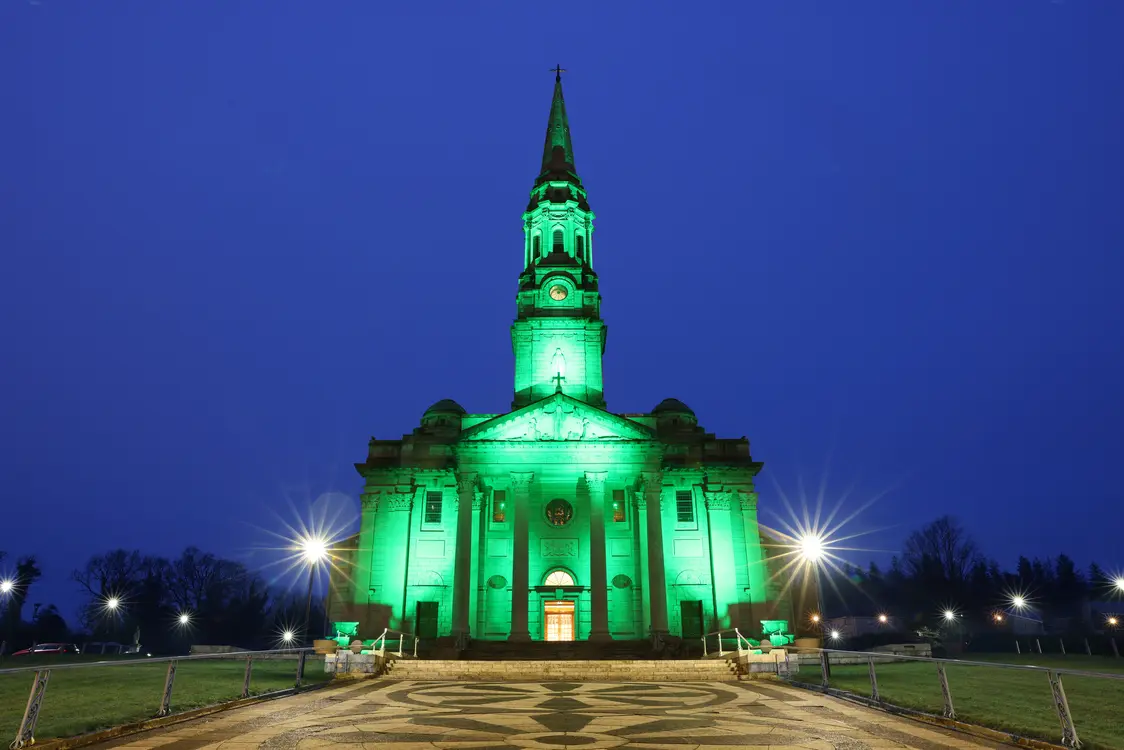 Cathedral of Saint Patrick and Saint Felim, Cavan - Illuminated in green light, a cathedral with a tall central spire and grand columned entrance, photographed at dusk under a deep blue sky. A patterned stone walkway leads to the front steps, with glowing streetlights and grassy lawns framing the symmetrical scene