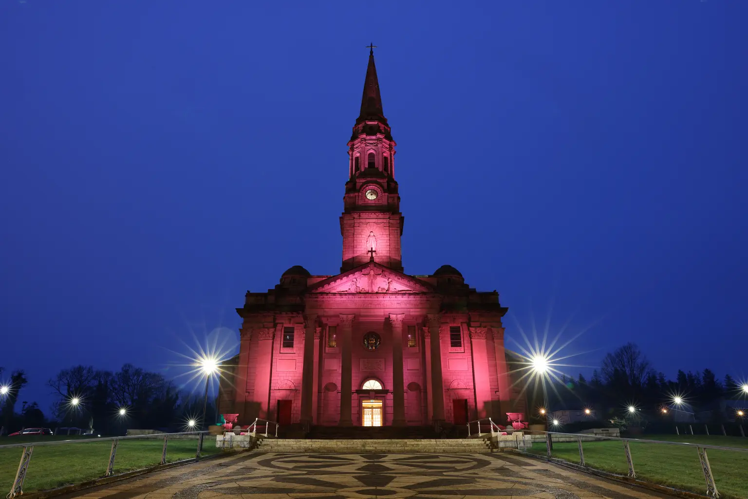 Cathedral of Saint Patrick and Saint Felim, Cavan - Illuminated in dark pink light, a cathedral with a tall central spire and grand columned entrance, photographed at dusk under a deep blue sky. A patterned stone walkway leads to the front steps, with glowing streetlights and grassy lawns framing the symmetrical scene