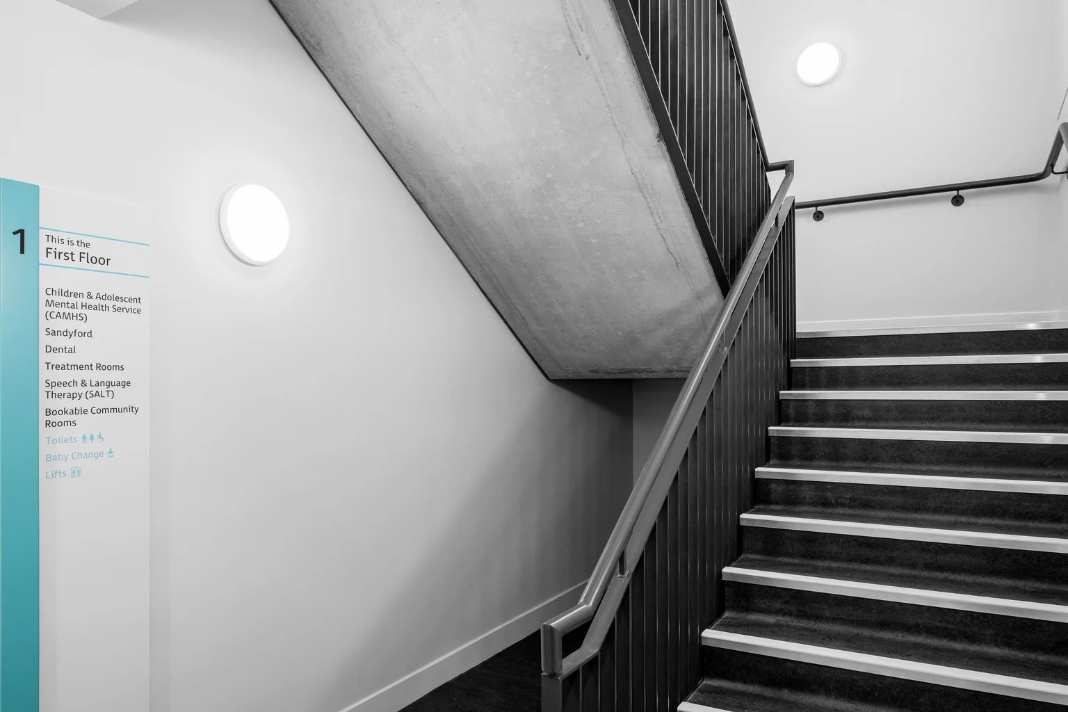 Een trappenhuis verlicht door ronde, aan de muur bevestigde ledlampen. Het minimalistische ontwerp heeft zwarte balustrades, betonnen elementen en duidelijke bewegwijzering voor een strakke en praktische uitstraling.