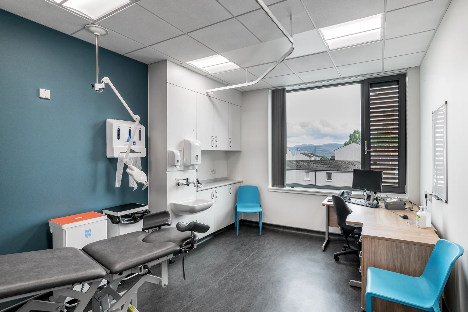 Een moderne medische onderzoeksruimte met een strak design, voorzien van een behandeltafel, een bureau met computer en felblauwe stoelen. De ruimte is goed verlicht met natuurlijk licht dat door een groot raam valt en aangevuld wordt met plafondverlichting, waardoor een nette en professionele sfeer ontstaat.