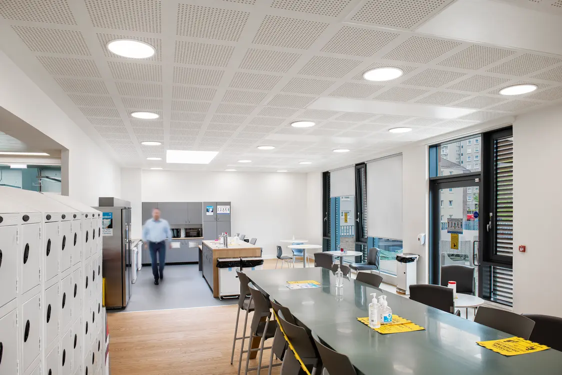 Une salle de pause lumineuse avec de longues tables, des chaises et des casiers sur le côté. À l’arrière-plan se trouve une kitchenette, et une personne traverse la pièce.