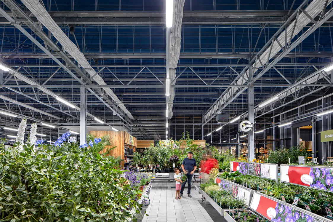 Ein moderner Gartenmarkt mit einer hohen Glas- und Stahlkonstruktion, die viel Tageslicht hineinlässt. Die Pflanzen und Blumen sind übersichtlich angeordnet, was eine angenehme Einkaufsatmosphäre schafft.