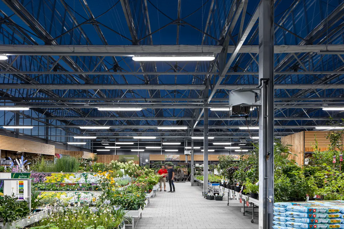 Ein moderner Gartenmarkt mit einer hohen Stahl-Glas-Konstruktion, die durch helle LED-Leuchten optimal ausgeleuchtet wird. Die Pflanzen sind übersichtlich angeordnet, und die offene Gestaltung schafft eine einladende Atmosphäre.