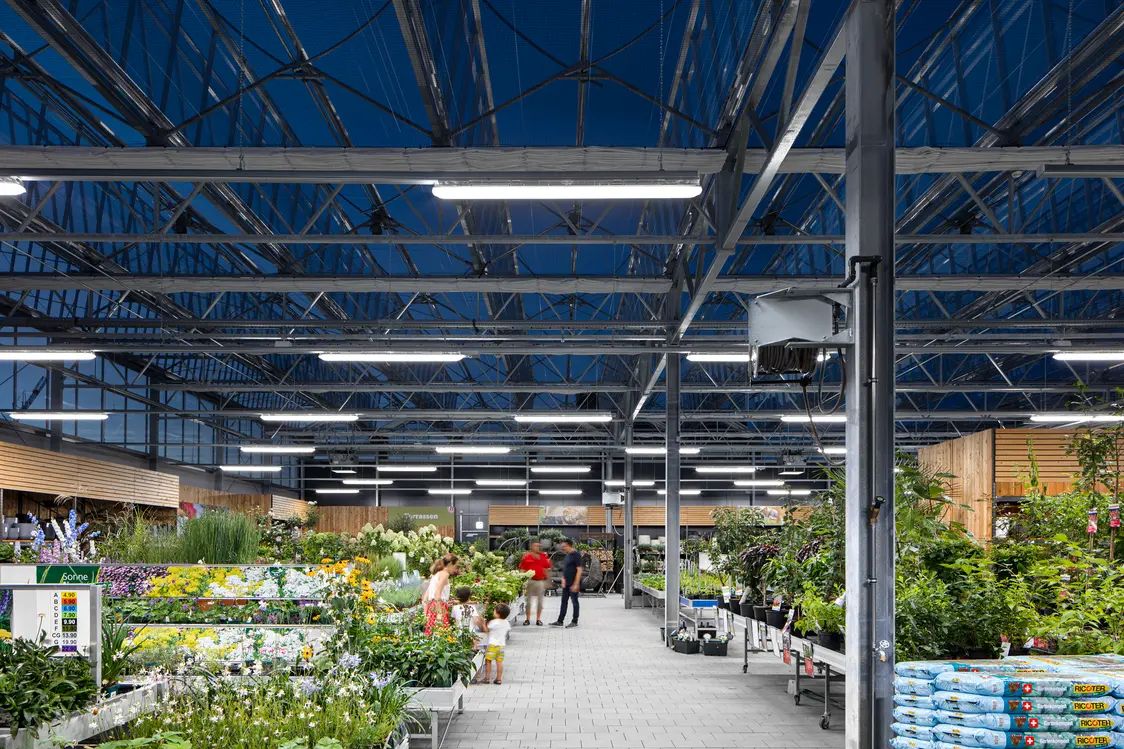 Ein moderner Gartenmarkt mit einer hohen Stahl-Glas-Konstruktion, die durch helle LED-Leuchten auch bei Dunkelheit optimal ausgeleuchtet ist. Die Pflanzen sind übersichtlich angeordnet und schaffen eine einladende Atmosphäre für Kunden.