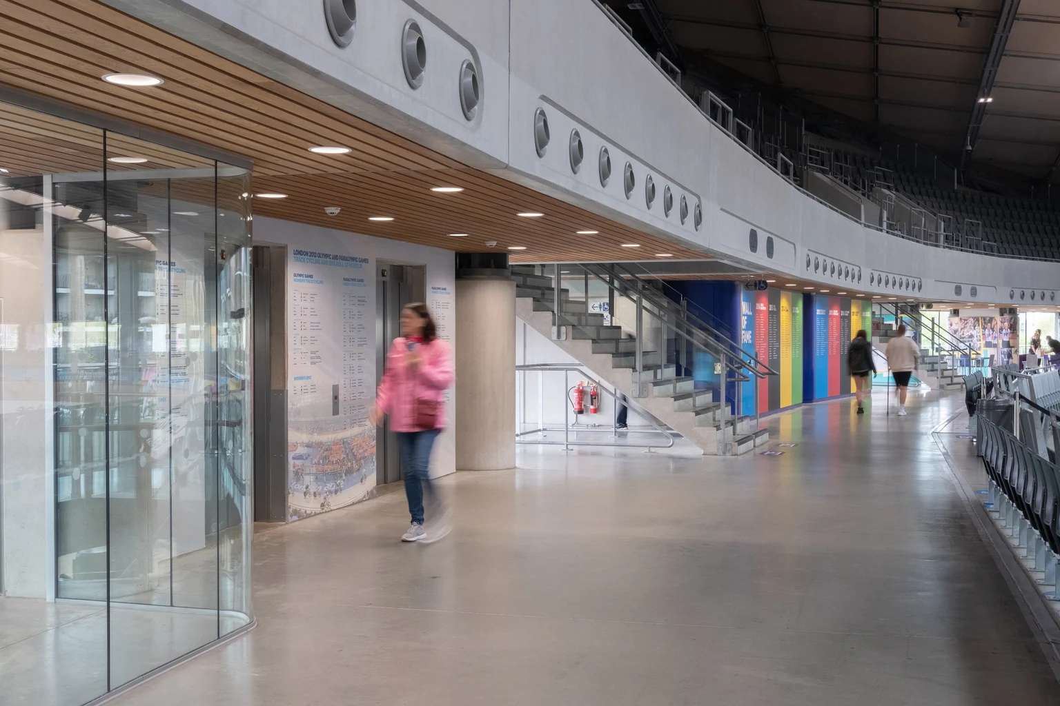 L'interno di un moderno velodromo presenta una spaziosa passerella con pareti divisorie in vetro, pannelli murali colorati e aree di seduta sopraelevate. Una persona passa sotto le luci brillanti del soffitto, aggiungendo a un'atmosfera vivace e aperta.