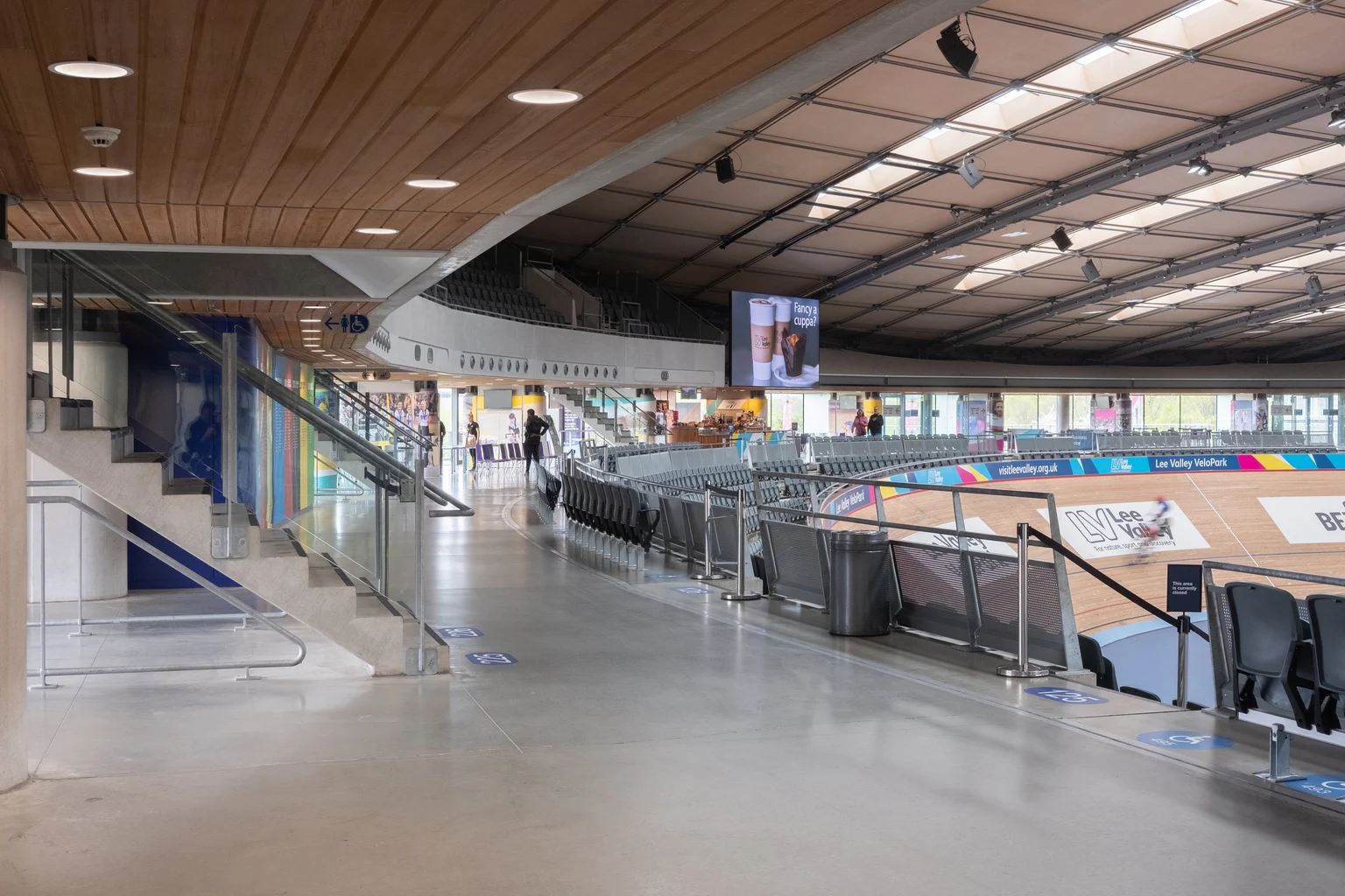 L'interno di un moderno velodromo con un soffitto curvo in legno e ampie aree di seduta che si affacciano sulla pista ciclabile. Ringhiere in vetro e luce naturale brillante dalle grandi finestre migliorano il design aperto e contemporaneo.