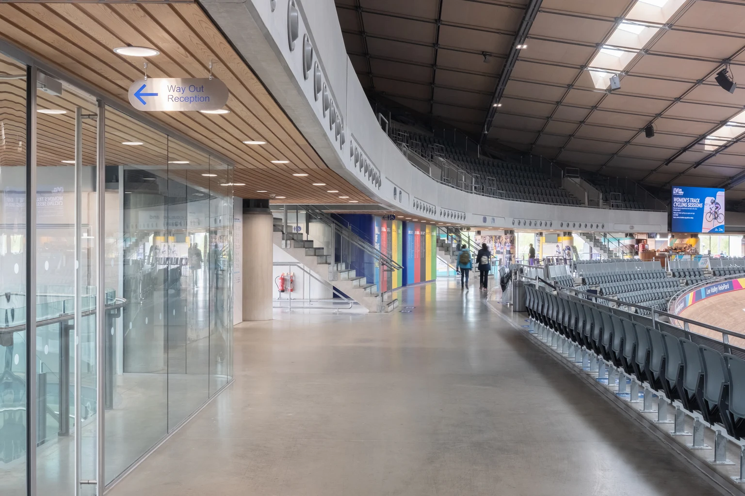 L'interno di un moderno velodromo con soffitto in legno curvo, pareti divisorie in vetro e file di posti a sedere che si affacciano sulla pista ciclabile. L'illuminazione brillante e i pannelli murali colorati creano un'atmosfera aperta e invitante.