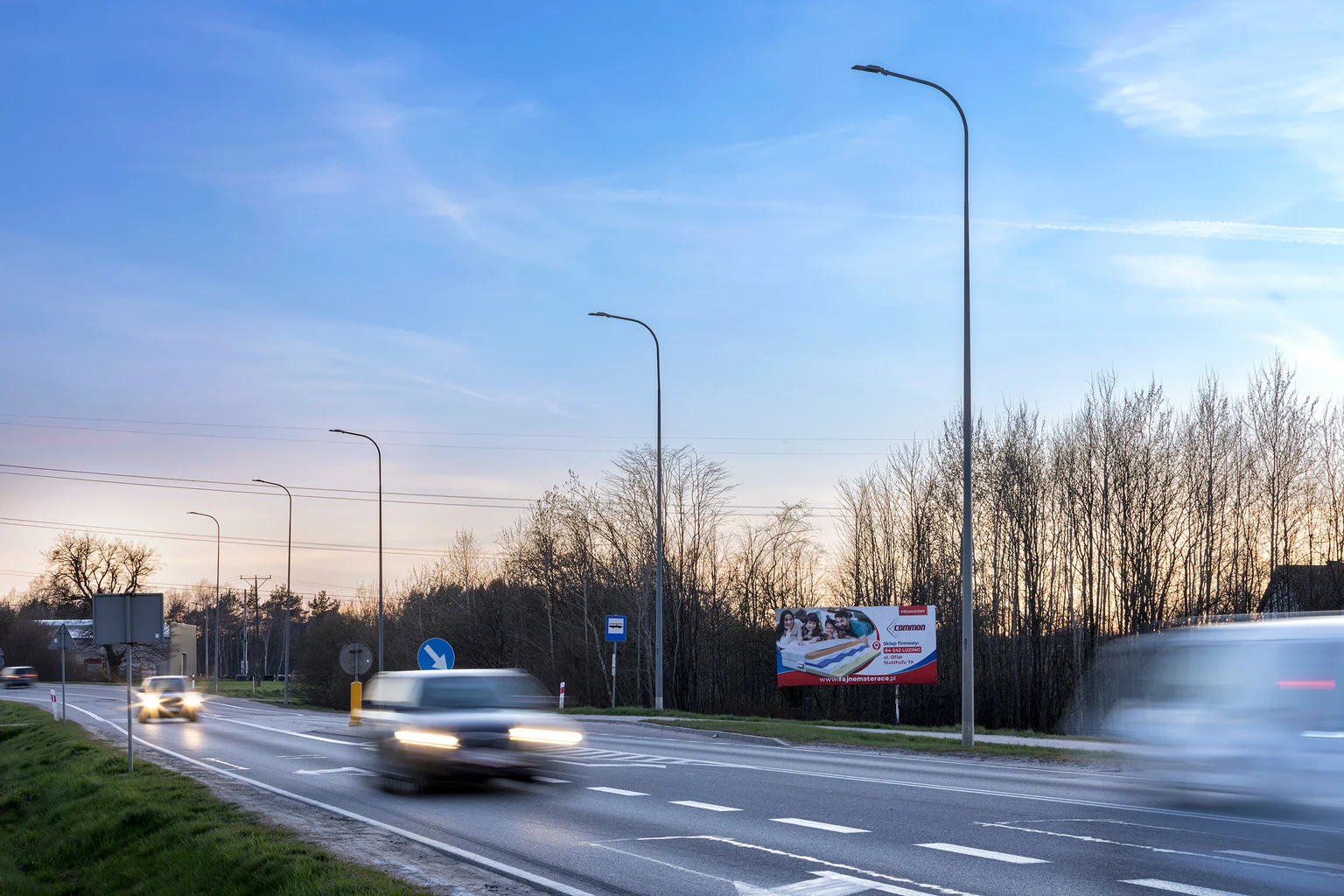 Eine Vorortstraße in der Abenddämmerung, beleuchtet von modernen Straßenlaternen mit vorbeifahrenden Autos. Im Hintergrund sind Bäume und eine bunte Werbetafel unter einem klaren Abendhimmel zu sehen.