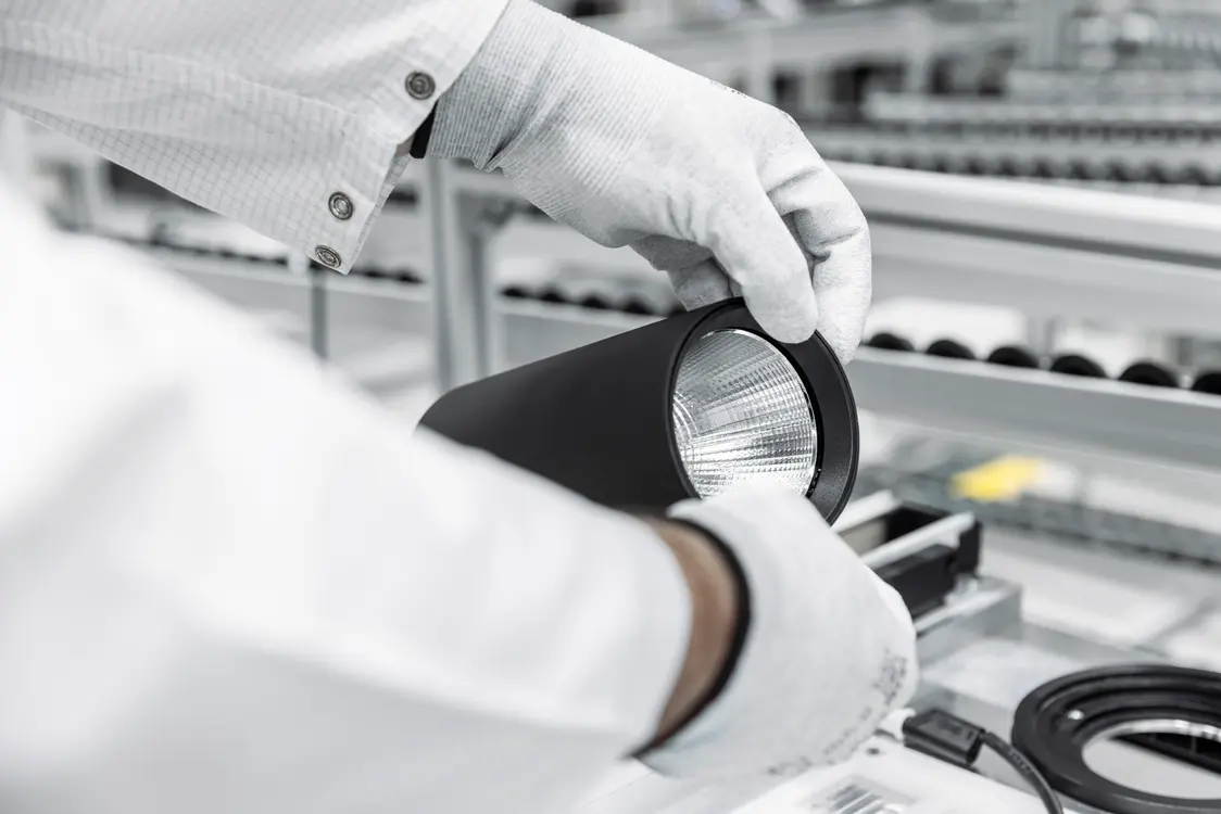 Close-up of a person wearing white gloves carefully holding a cylindrical optical component in a clean, industrial or laboratory setting, with shelves of similar parts blurred in the background.