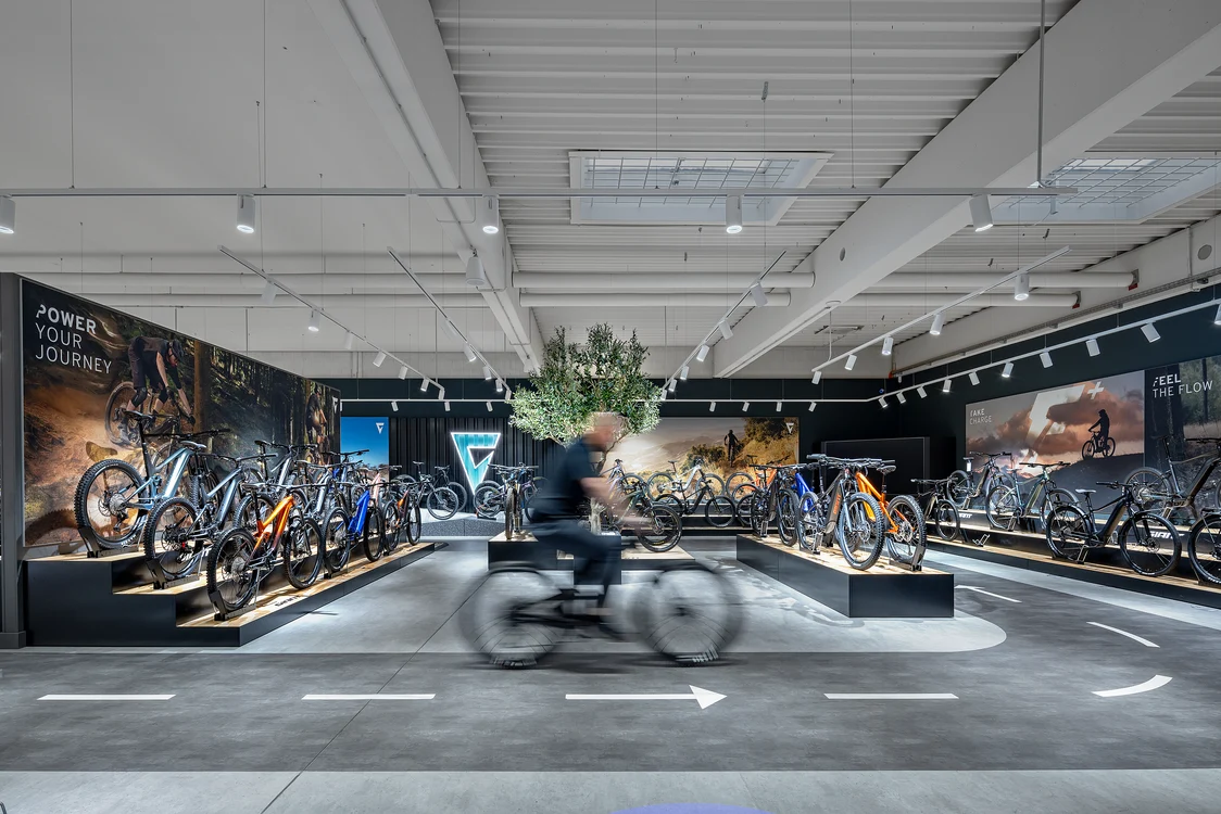  Ausstellungsraum mit zahlreichen Fahrrädern und einem Kunden auf dem Fahrrad, die Beleuchtung besteht aus gleichmäßig angeordneten Deckenstrahlern, die die Produkte optimal in Szene setzen.