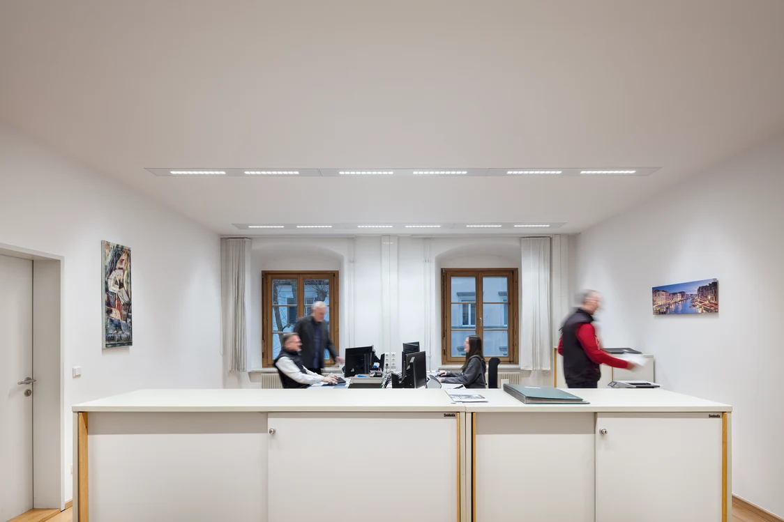 Ufficio luminoso con pareti bianche, postazioni di lavoro moderne e luci LED lineari sul soffitto. Tre persone lavorano alle scrivanie di fronte a grandi finestre, mentre alle pareti sono appesi dei quadri.
