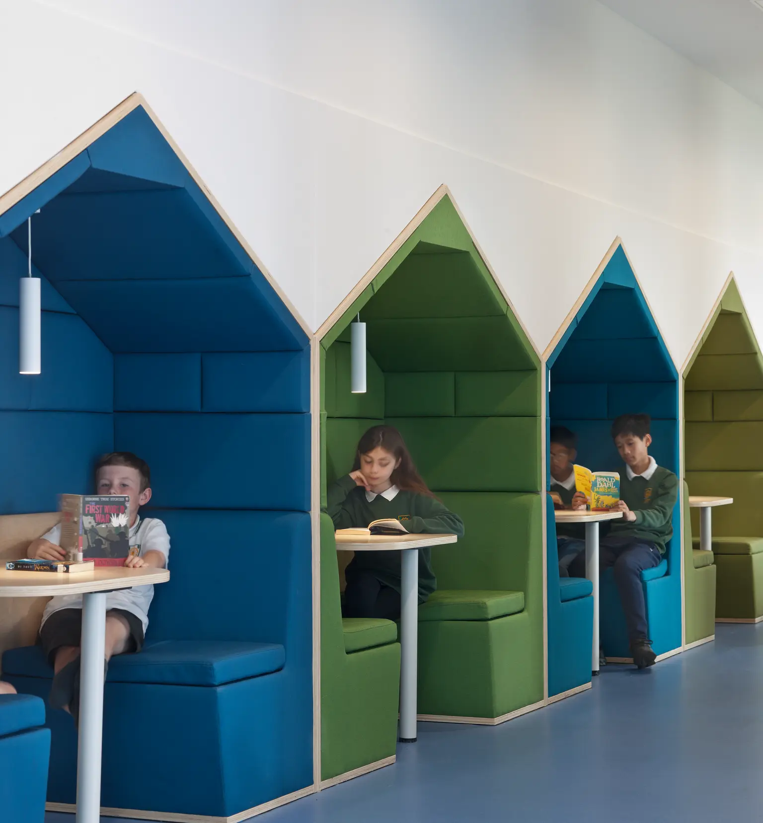 Milltimber Primary School, Aberdeen - Several young children sit reading books in colourful, house-shaped study modern booths in a school learning space. The booths are upholstered in blue and green fabric, with built-in benches, small tables, and hanging pendant lights.