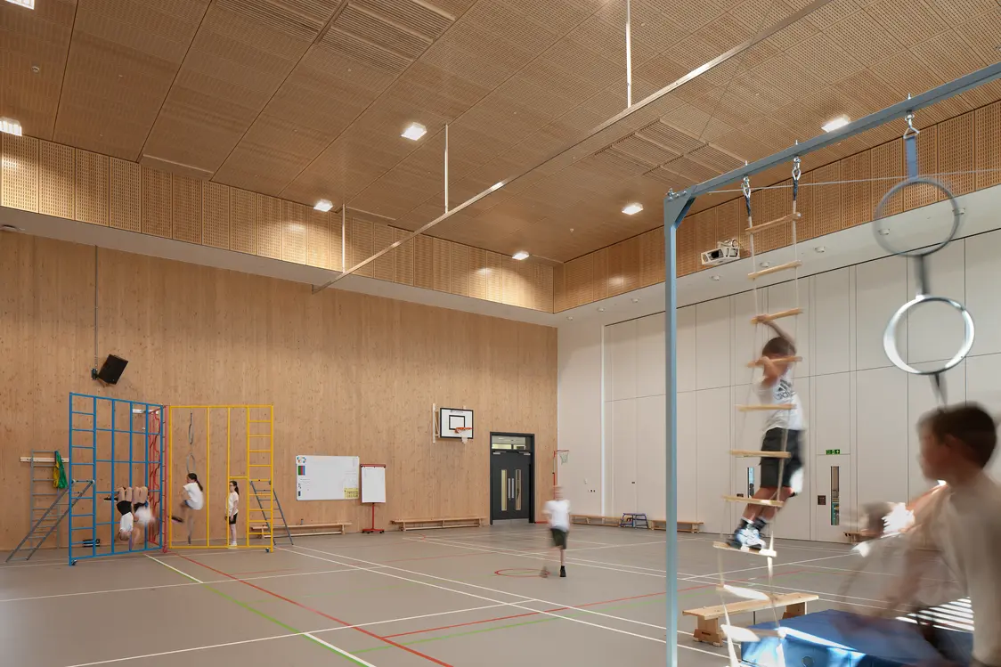 Milltimber Primary School, Aberdeen - Large school sports hall with timber-clad walls and ceiling, featuring climbing frames, gymnastic rings and suspended play equipment. Several children are actively using the equipment and moving across the marked indoor court, creating a dynamic physical education environment
