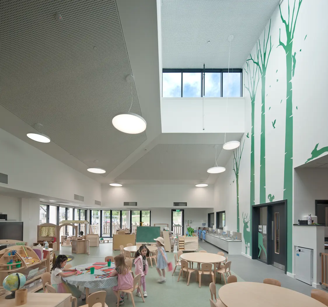 Milltimber Primary School, Aberdeen – Bright children's learning and play area with wooden furniture, round activity tables and learning stations. Several young children are engaged in play and creative activities throughout the open-plan space, which features large windows, high ceilings, and a wall decorated with green tree graphics