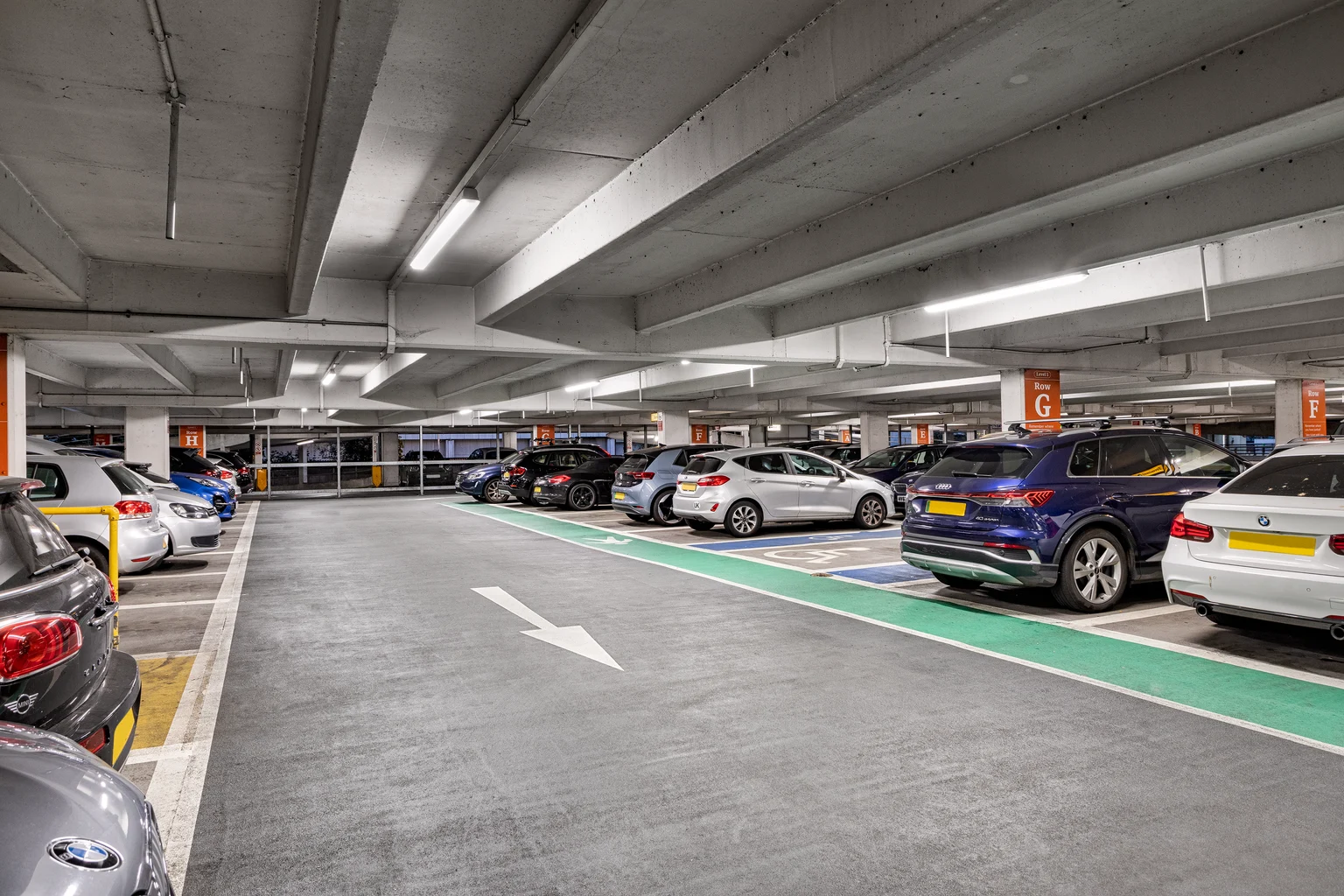 Ein hell beleuchtetes mehrstöckiges Parkhaus mit markierten Parkplätzen, Grünzonen für Elektrofahrzeuge und auf beiden Seiten geparkten Autos. Zur einfachen Navigation sind die Abschnitte mit „G“ beschildert.