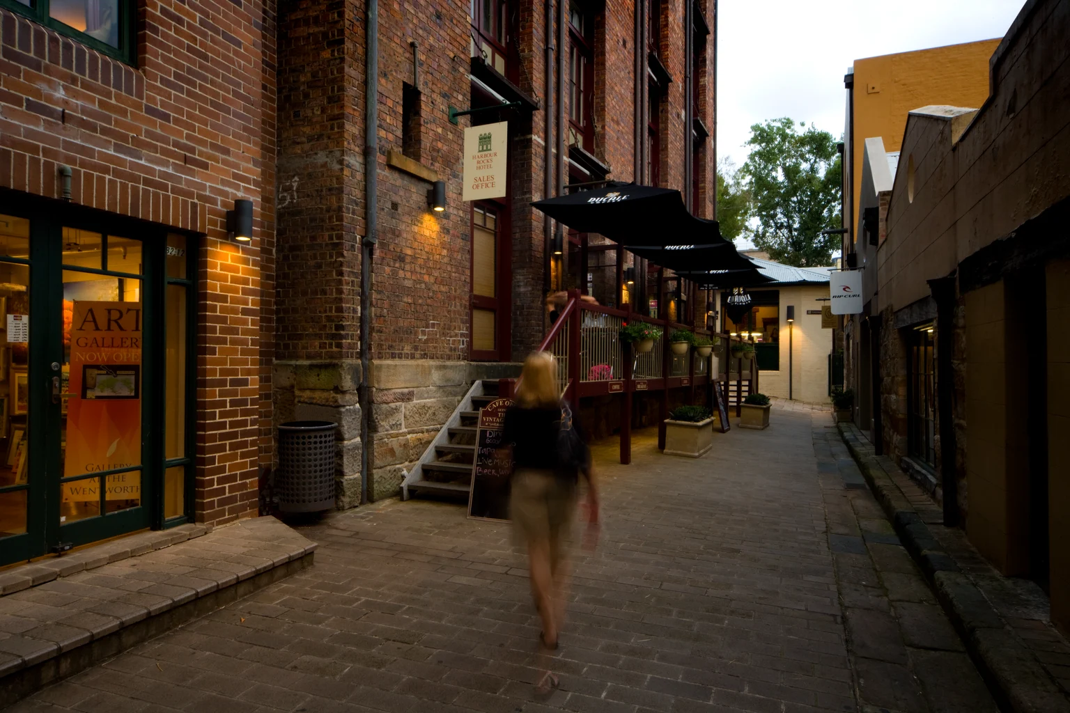 Cesar Amp éclairant doucement l'entrée d'une galerie d'art tandis qu'une dame marche dans une rue pavée