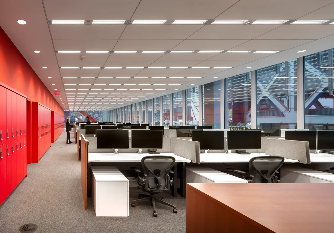 Großraumbüro mit modernen Arbeitsplätzen, ausgestattet mit Schreibtischen, Monitoren und ergonomischen Bürostühlen, sowie einer Fensterfront, die viel Tageslicht hereinlässt. Im Hintergrund sind zwei Personen in einer Unterhaltung zu sehen.