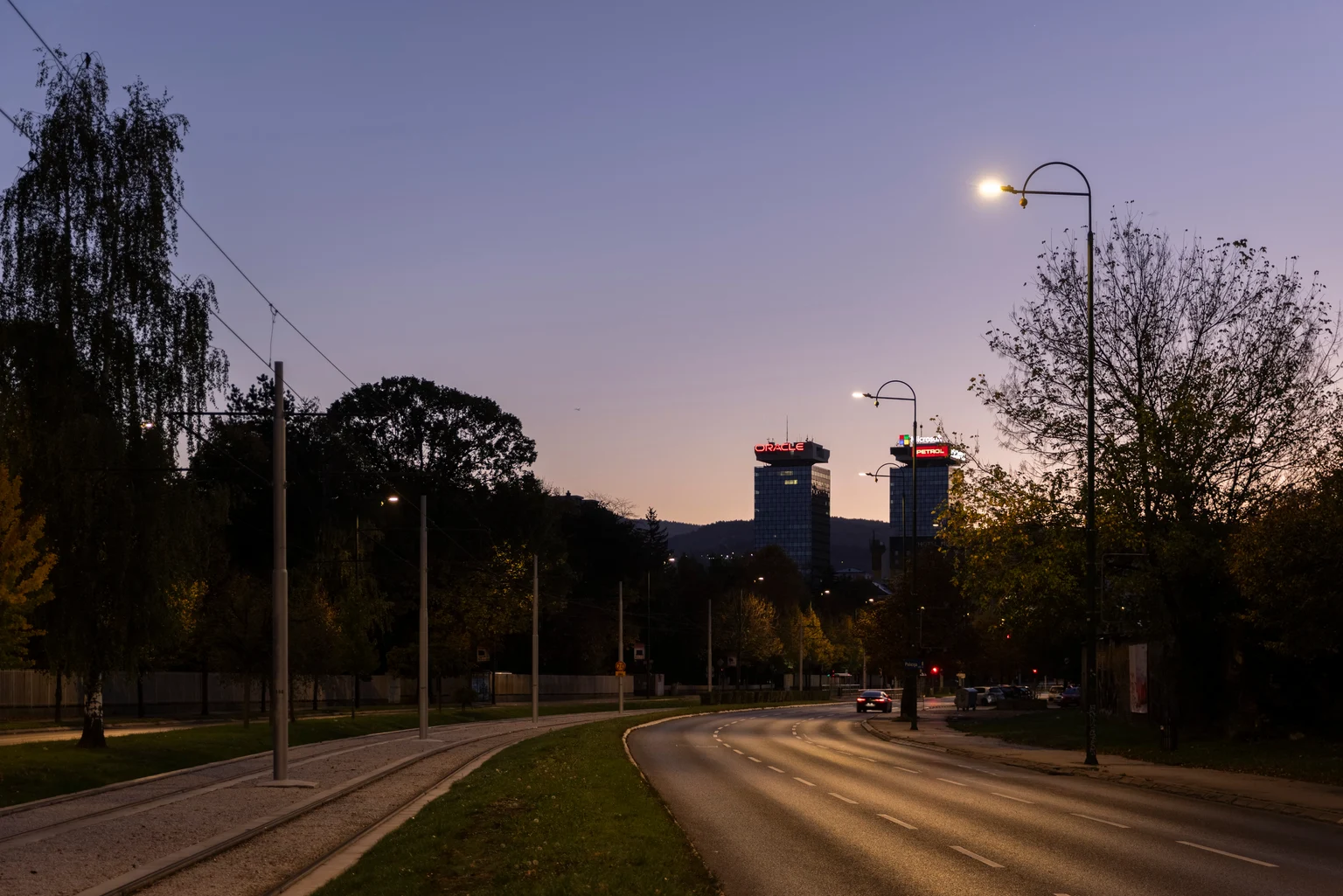 Eine Vorortstraße in der Abenddämmerung mit gebogenen Straßenleuchten und Bäumen auf beiden Seiten. Im Hintergrund befinden sich zwei hohe Gebäude mit rot beleuchteten Dächern, die in den Abendhimmel ragen.