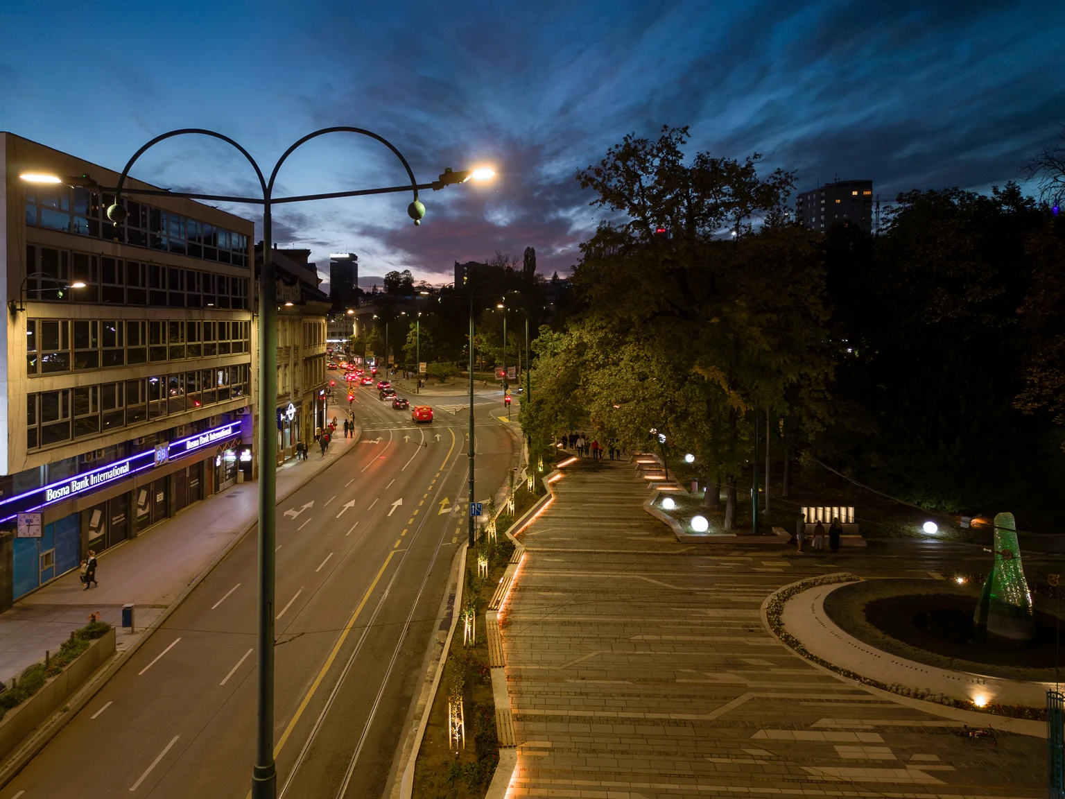 Eine Stadtstraße in der Abenddämmerung mit modernen gebogenen Straßenleuchten, die die Straße und den Fußgängerweg beleuchten. Auf der linken Seite bildet ein hell erleuchtetes Gebäude einen Kontrast zu Bäumen und einer Parkanlage auf der rechten Seite unter einem dramatischen Abendhimmel.