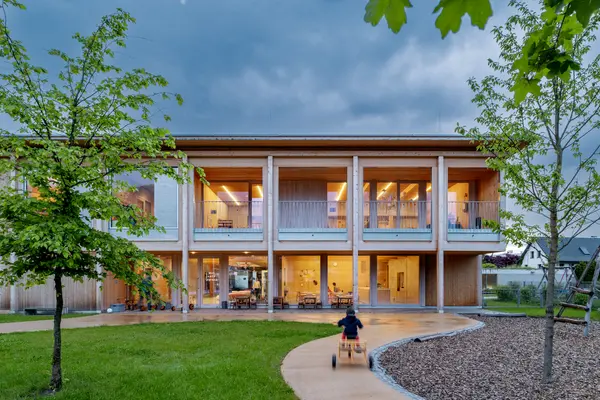  Maison d'enfants à deux étages avec une façade en bois, de grandes baies vitrées et un éclairage d'intérieur chaleureux. L'architecture adaptée aux enfants intègre des matériaux naturels avec une conception d'éclairage invitante.