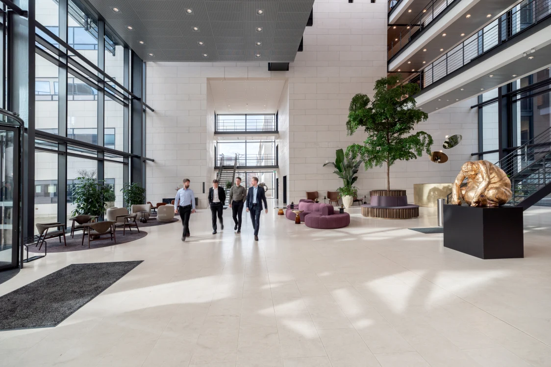Spacious, modern entrance area with tall glass fronts, light stone floors, and elegant seating areas. Several individuals walk through the light-filled lobby, which is adorned with plants and art pieces.