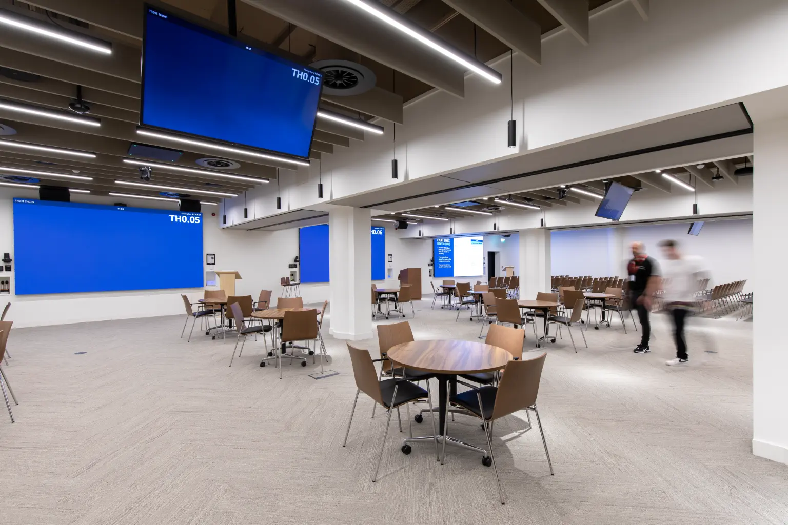 Capital One, Nottingham - 2 people walking through a flexible meeting and event space with round tables, chairs, large display screens, and AV equipment