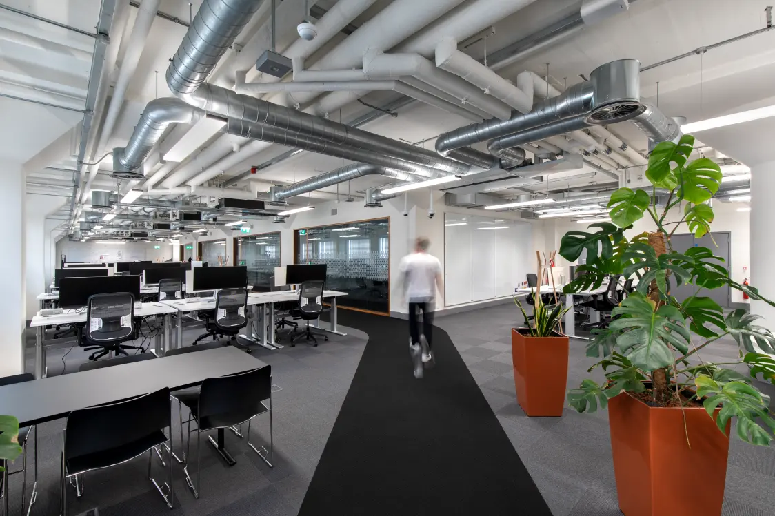 Capital One, Nottingham - A person walks through an open plan office with desks, ergonomic chairs, indoor plants, and exposed ceiling ductwork in a modern workspace