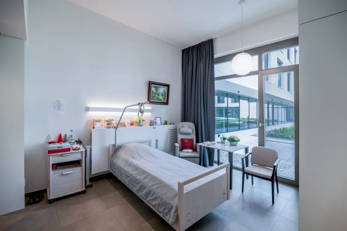 Une chambre de patient confortable, aménagée avec un lit simple, une table et des chaises. Une suspension LED diffuse une lumière chaude, complétée par une lampe de luminothérapie installée près du lit. De grandes fenêtres apportent un maximum de lumière naturelle, contribuant à une ambiance apaisante.