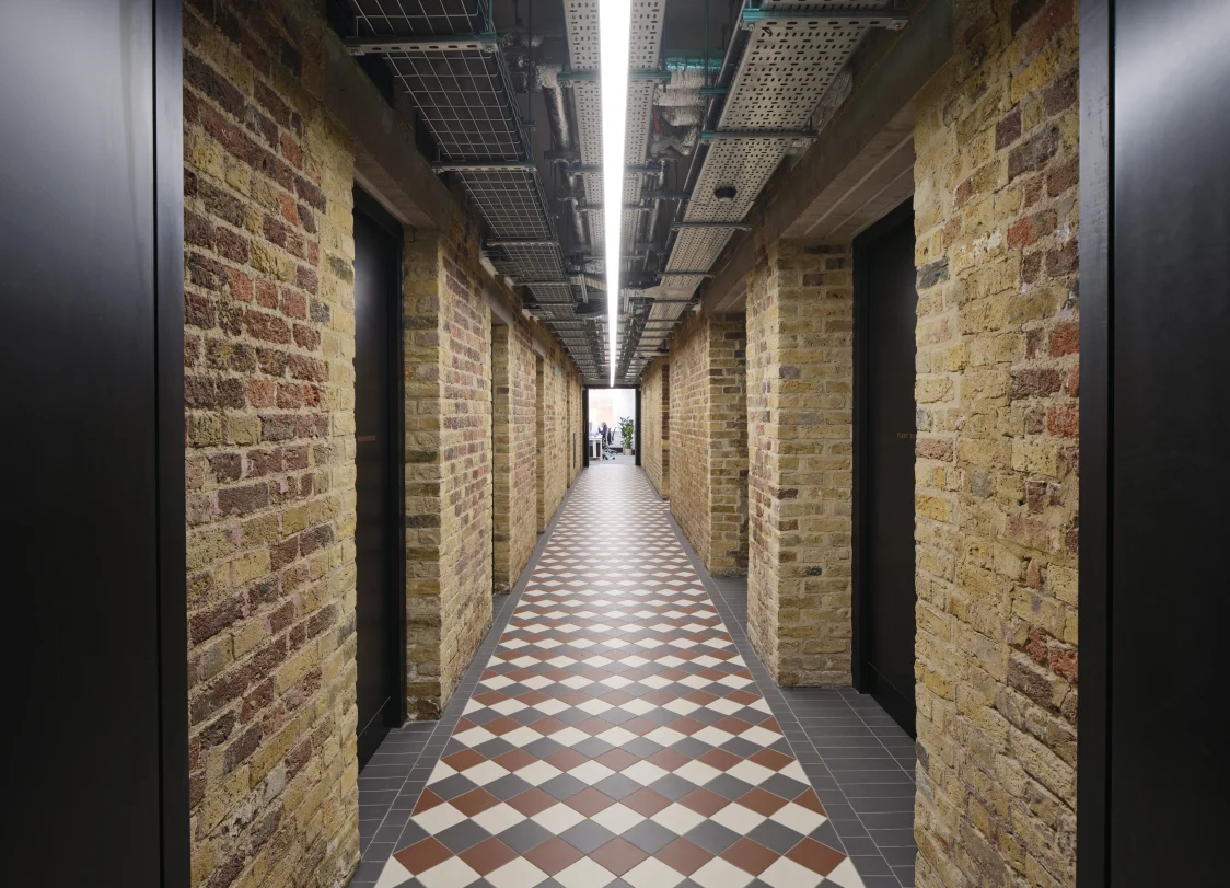 Narrow corridor with exposed brickwork, historic floor pattern, and open technical ceiling. A linear lighting solution provides uniform illumination and highlights the depth of the space.