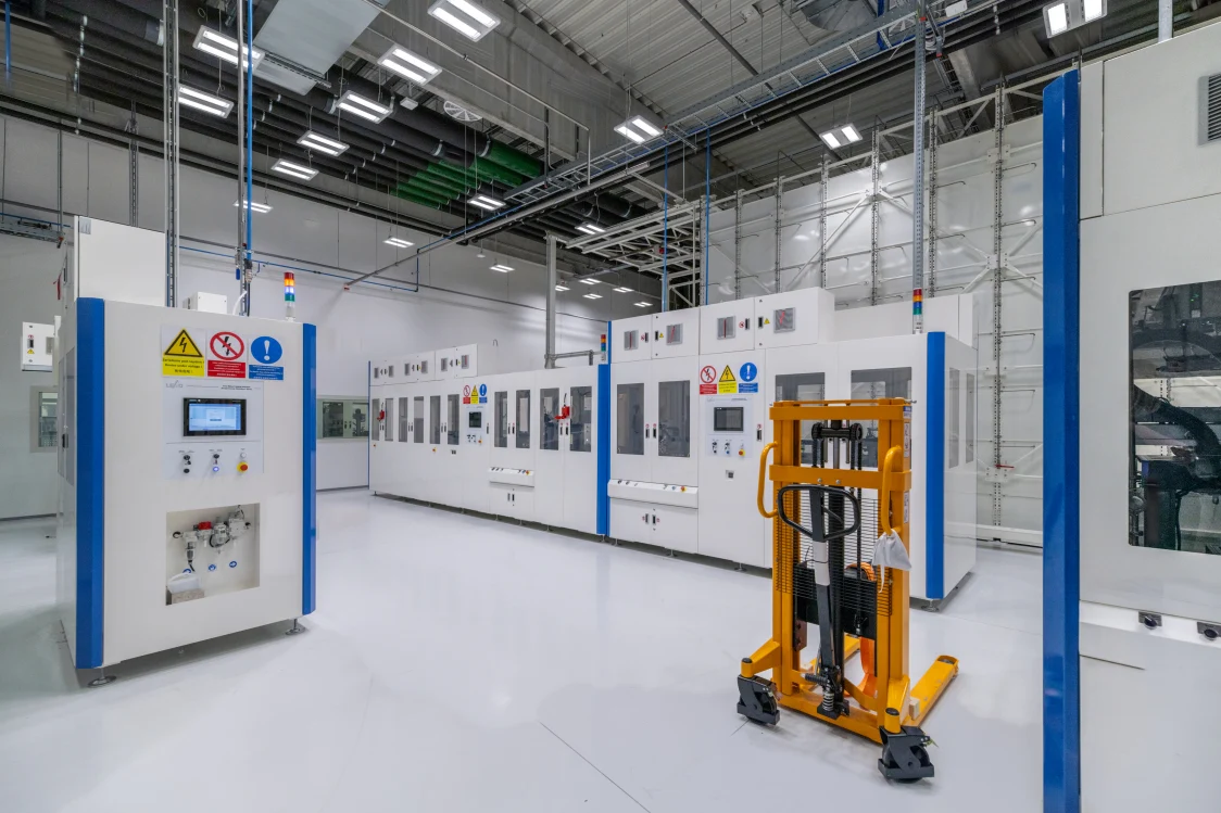 An industrial production hall with white machines and blue columns, illuminated by bright LED lights on the ceiling. A yellow pallet truck is in the foreground, while the clear lighting highlights the technical details.