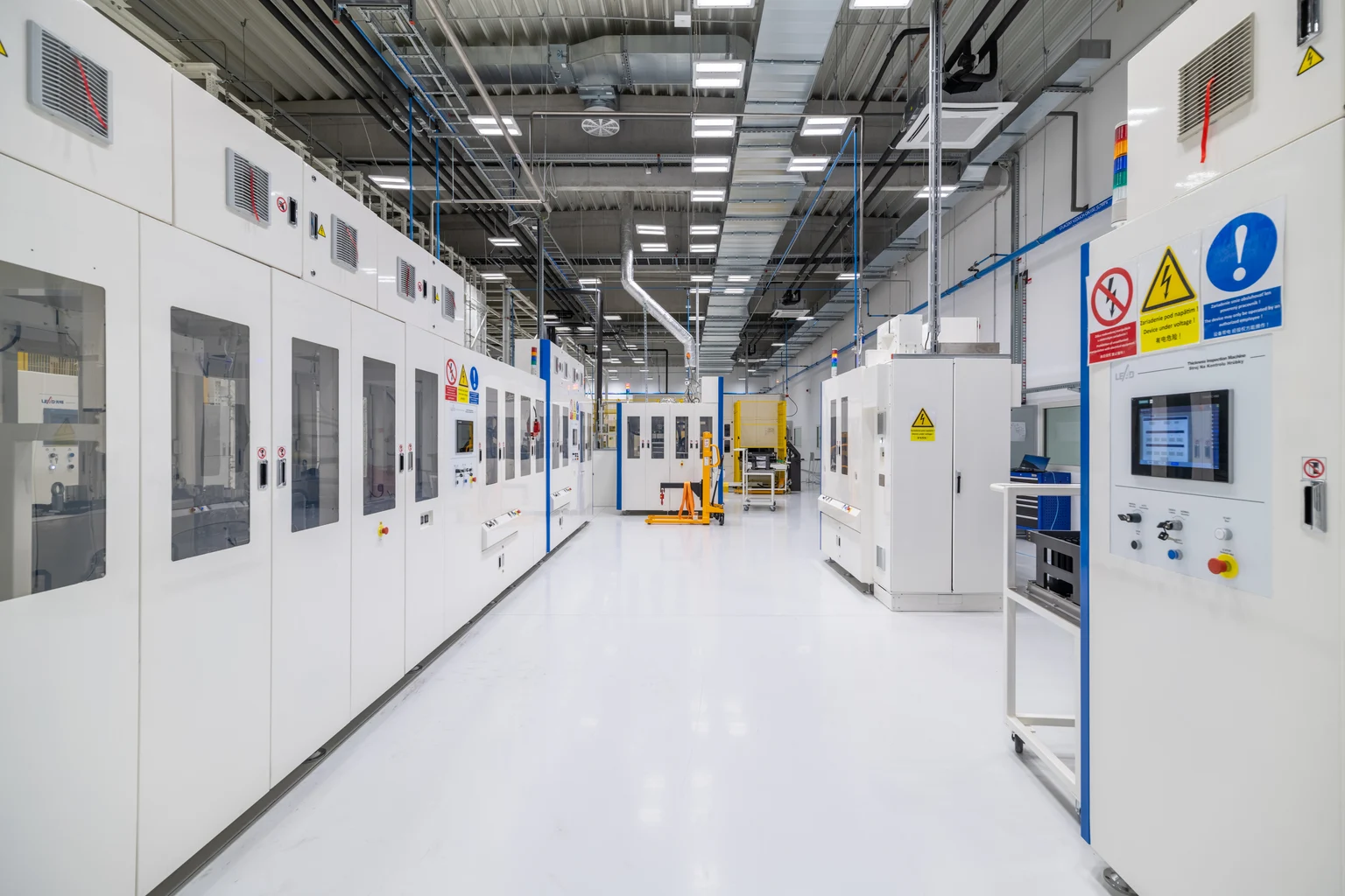 Un hall de production industriel avec des machines blanches et des colonnes bleues, éclairé par des luminaires LED brillants au plafond. Un transpalette jaune est placé au premier plan, tandis qu’une lumière claire met en valeur les détails techniques.