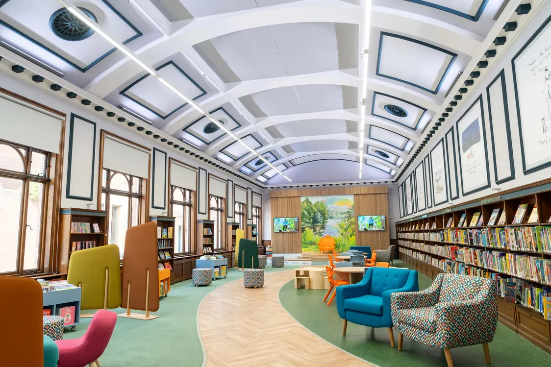 Une salle de bibliothèque lumineuse avec un plafond voûté blanc, éclairée par de longues réglettes LED. Les étagères à livres et les sièges colorés créent une atmosphère accueillante, tandis qu’une zone d’écran est visible en arrière-plan.