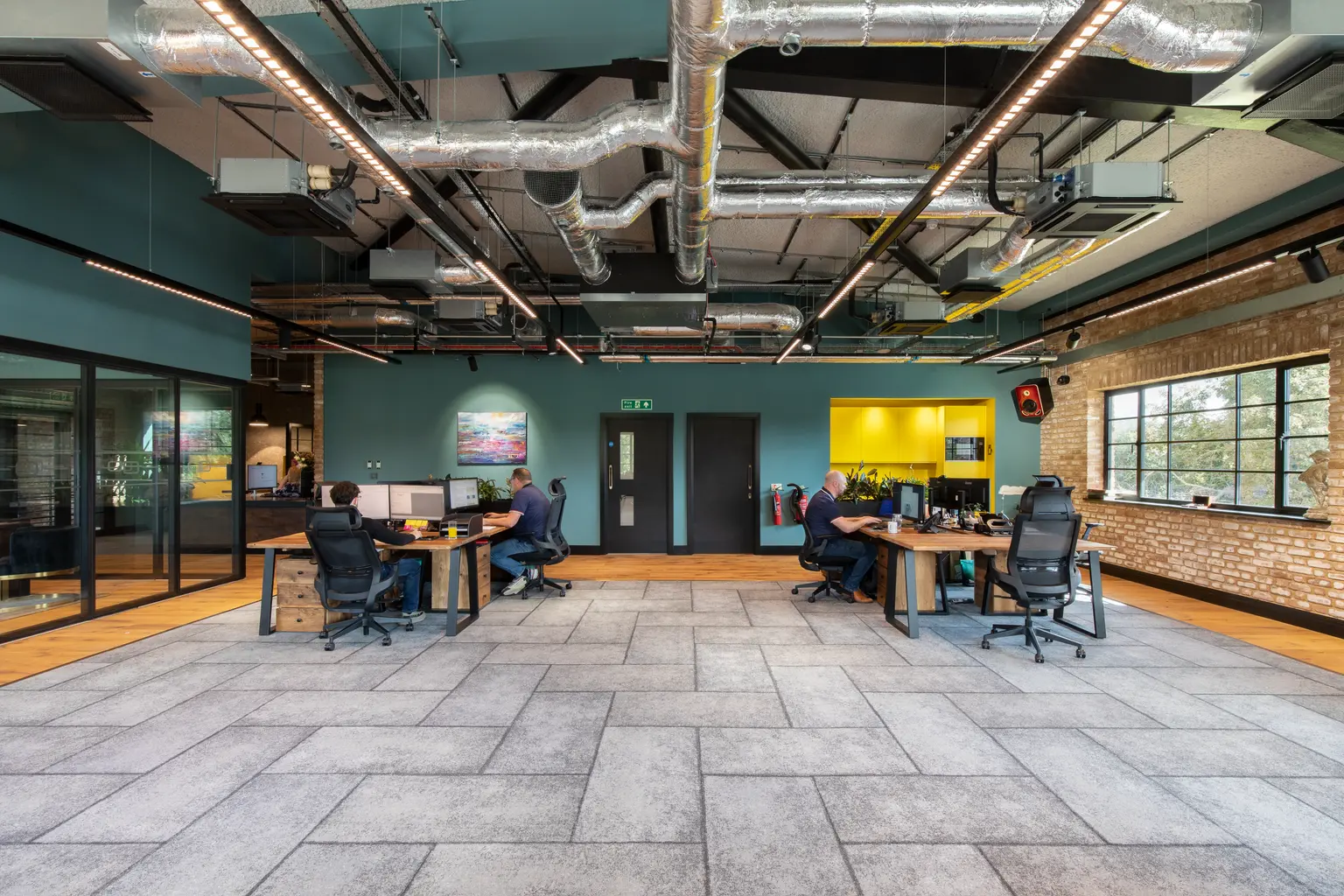 Oscar Acoustics, Rochester, Kent - Modern open-plan office with grey tiled floor, exposed ductwork, wooden desks, glass meeting rooms, teal walls, a yellow kitchenette, and large windows with natural light