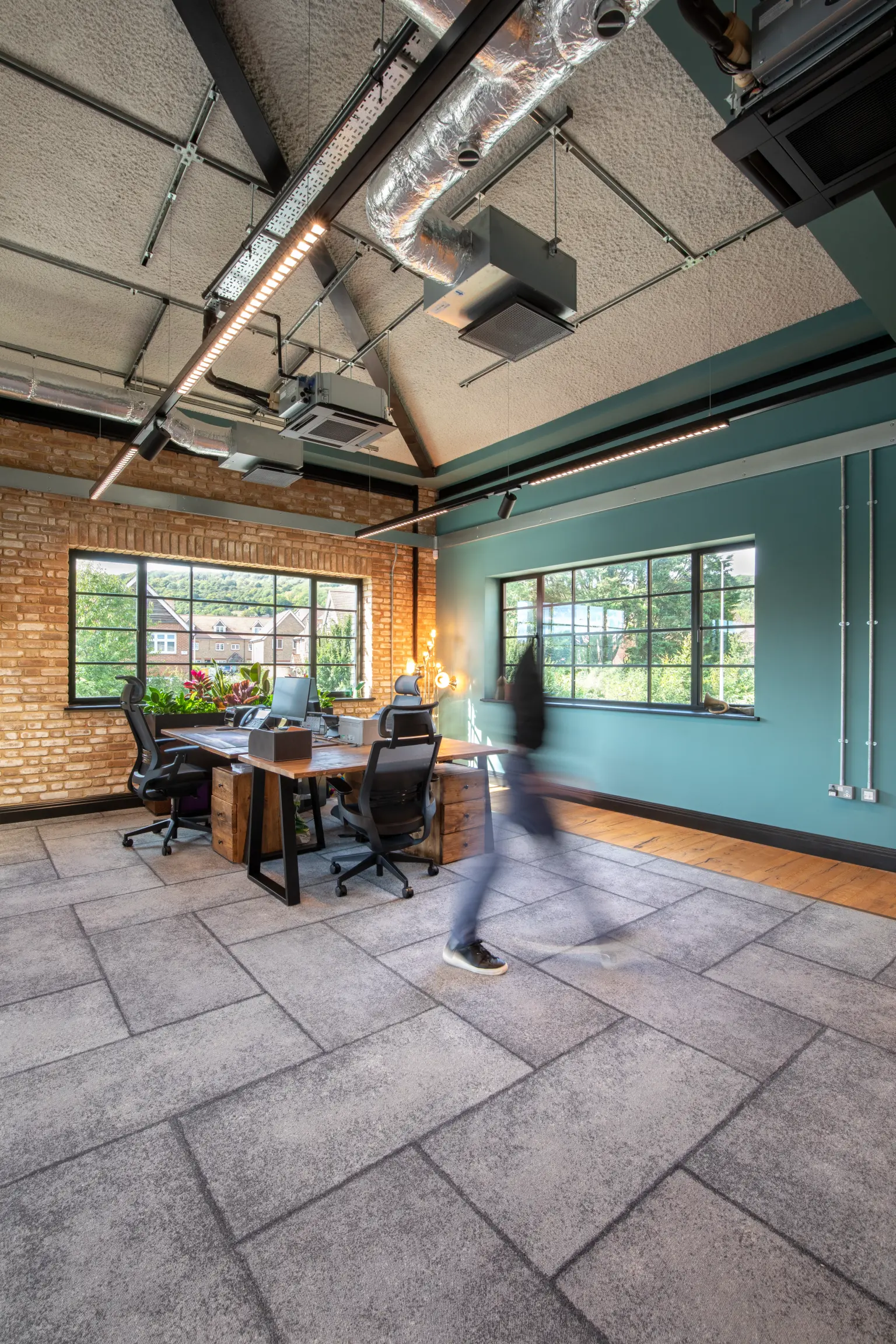Oscar Acoustics, Rochester, Kent - Contemporary open plan office with acoustic ceiling panels, exposed ceiling ducts, teal feature walls, wooden desks, large windows, and a brick accent wall. A blurred figure walks across the grey tiled floor.