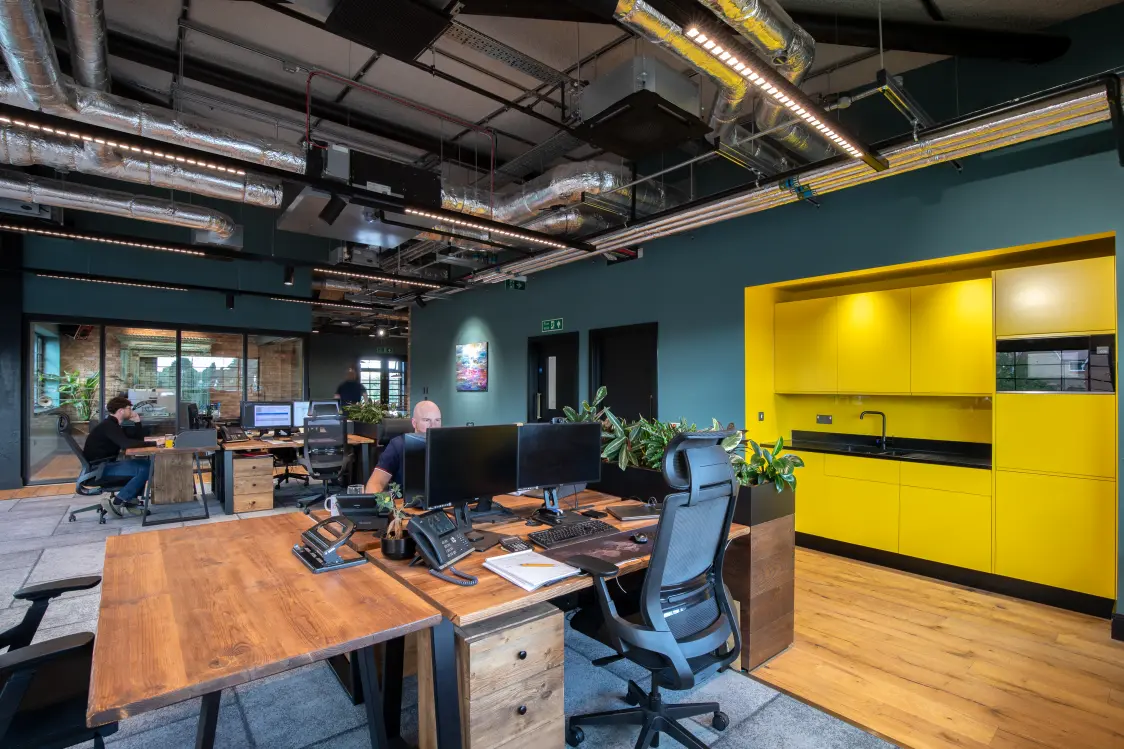 Oscar Acoustics, Rochester, Kent - Open-plan modern office with wooden desks, ergonomic chairs, and multiple computer workstations. Several people are working at their desks, while an industrial-style ceiling with exposed ductwork and linear lighting spans overhead. A bright yellow kitchenette contrasts with the dark teal walls, and glass-partitioned meeting rooms, indoor plants, and wood flooring create a contemporary, collaborative workspace