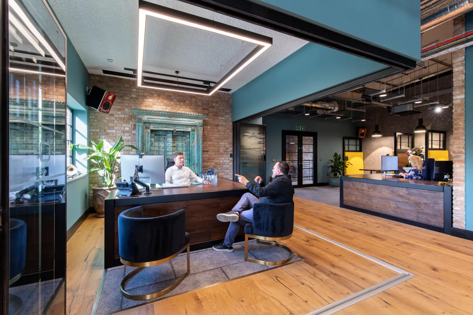 Oscar Acoustics, Rochester, Kent - Modern open plan office featuring wood flooring, exposed brick and teal walls, acoustic ceiling treatments, and contemporary workstations. Two people are seated in a meeting area in the foreground, with additional staff working at desks in the background