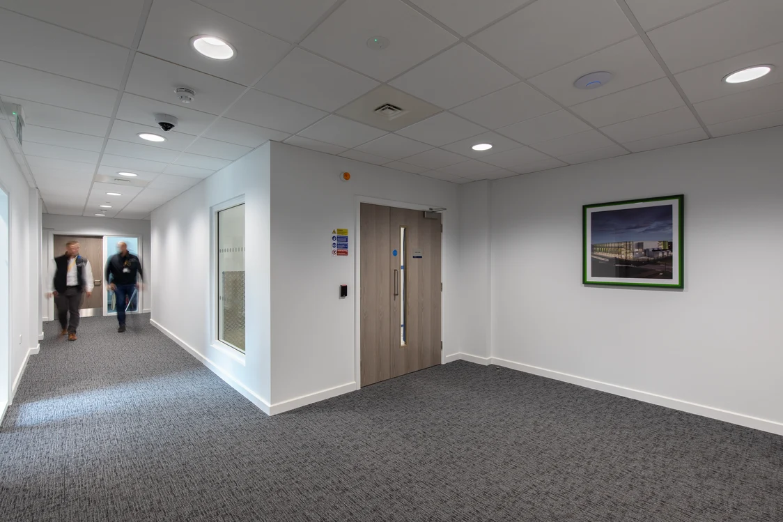 A bright office corridor with grey carpet and white walls with a picture hanging on them. Two people go towards a wooden door at the end of the corridor.