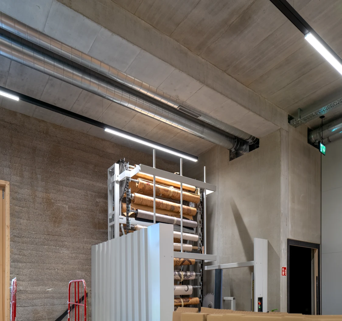 Un espace de stockage moderne avec murs en béton brut et une grande machine portant plusieurs rouleaux de matière. L’ensemble est propre et parfaitement éclairé.