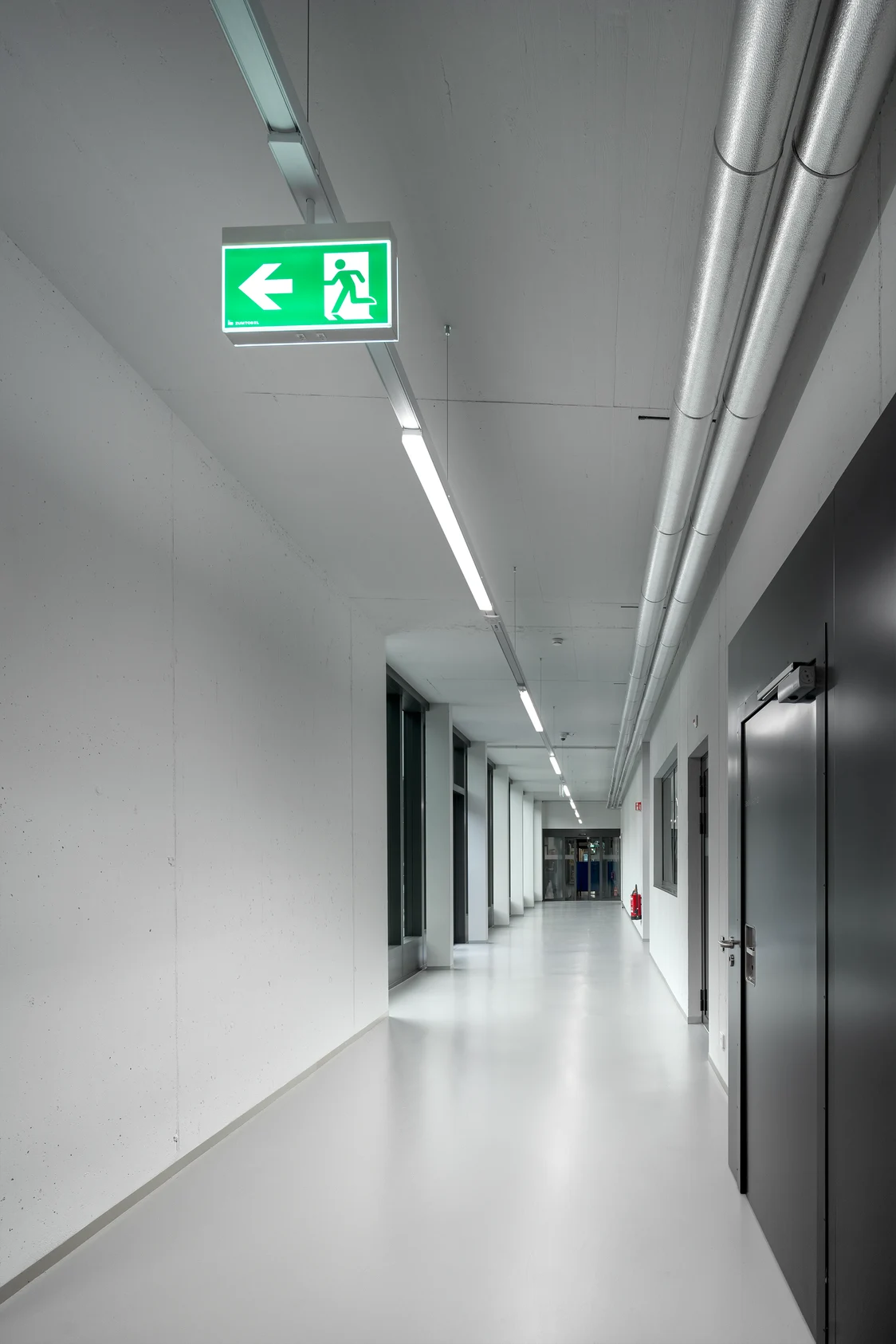 Un couloir blanc dans le site industriel. Un panneau de sortie de secours est suspendu en haut à gauche du plafond.