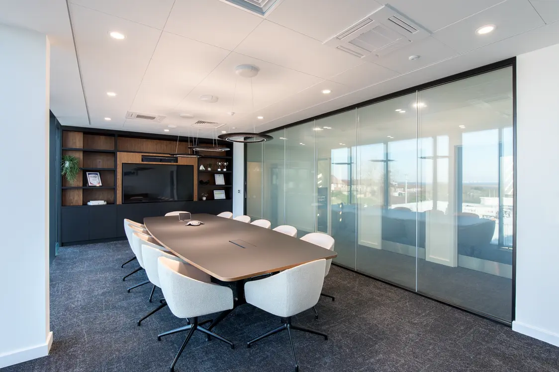 MEMS Power Generation Headquarters, Gillingham - Modern conference room with a large oval meeting table surrounded by eight upholstered chairs, a wall-mounted display set within dark wood shelving, and floor-to-ceiling glass partition walls overlooking an office exterior. Bright recessed lighting, grey carpet flooring, and a clean, contemporary design create a professional meeting space