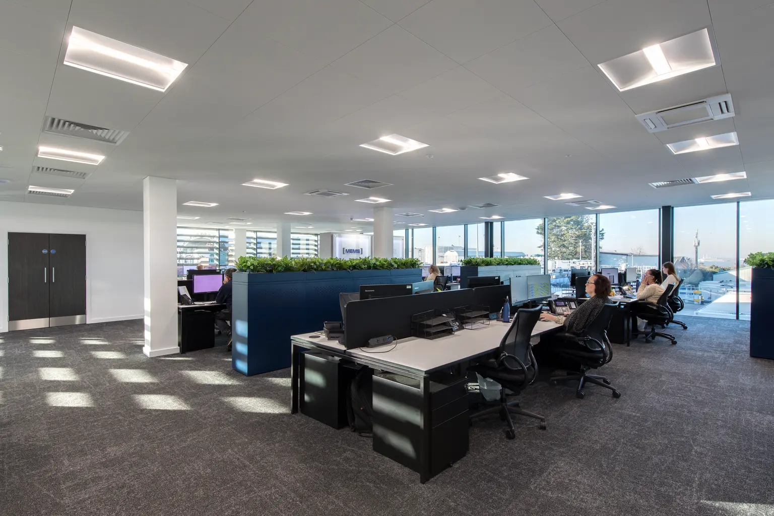 MEMS Power Generation headquarters, Gillingham - Open-plan office with rows of workstations, computer monitors and ergonomic chairs arranged around blue partition walls topped with greenery. Large floor-to-ceiling windows provide natural light across the modern workspace, where several employees are working at desksModern Open plan